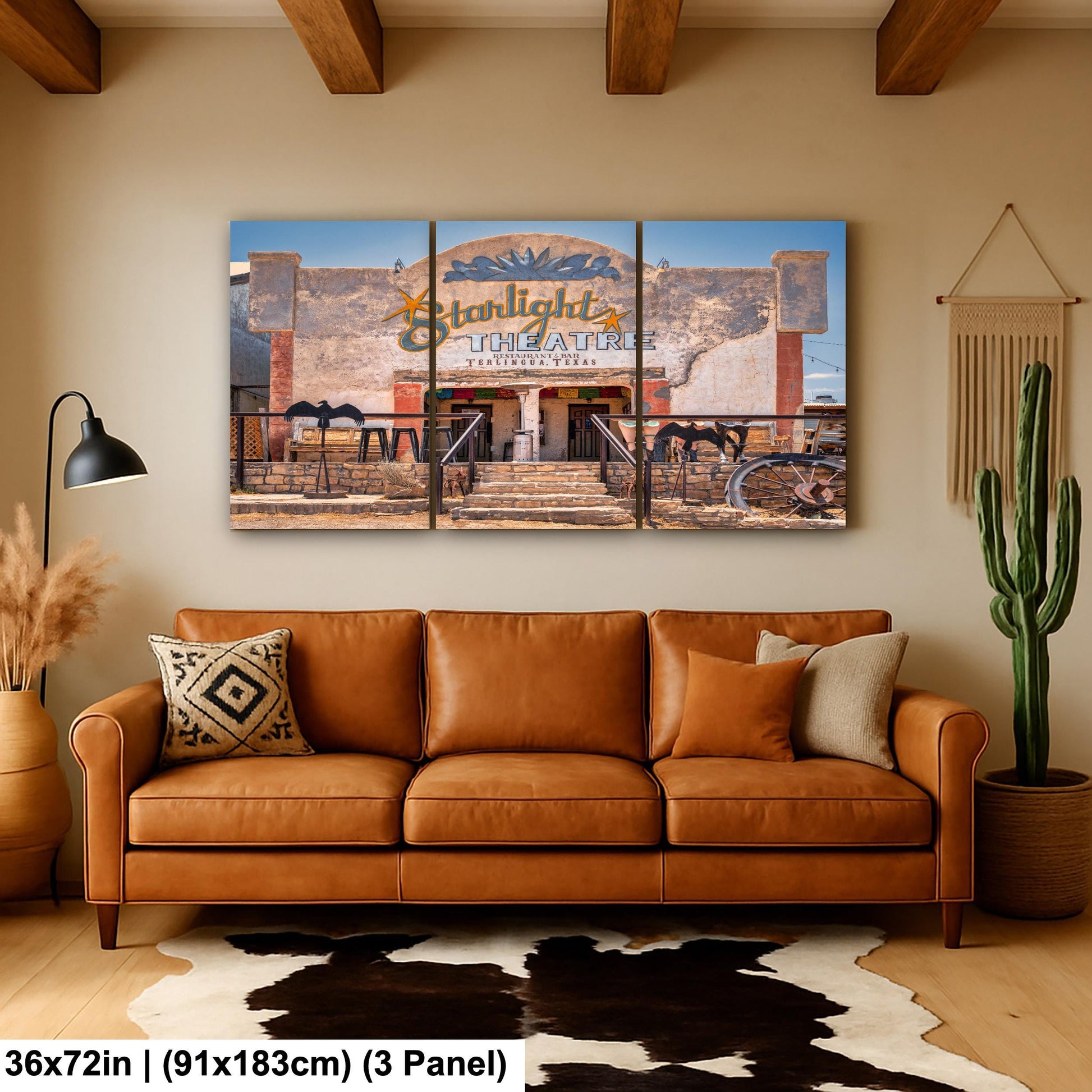 a large, three-panel print of a desert-like scene with a theater building in the background. The print is mounted on a wall above a brown leather couch, which is positioned in front of a cowhide rug. The overall setting appears to be a cozy, rustic living room.