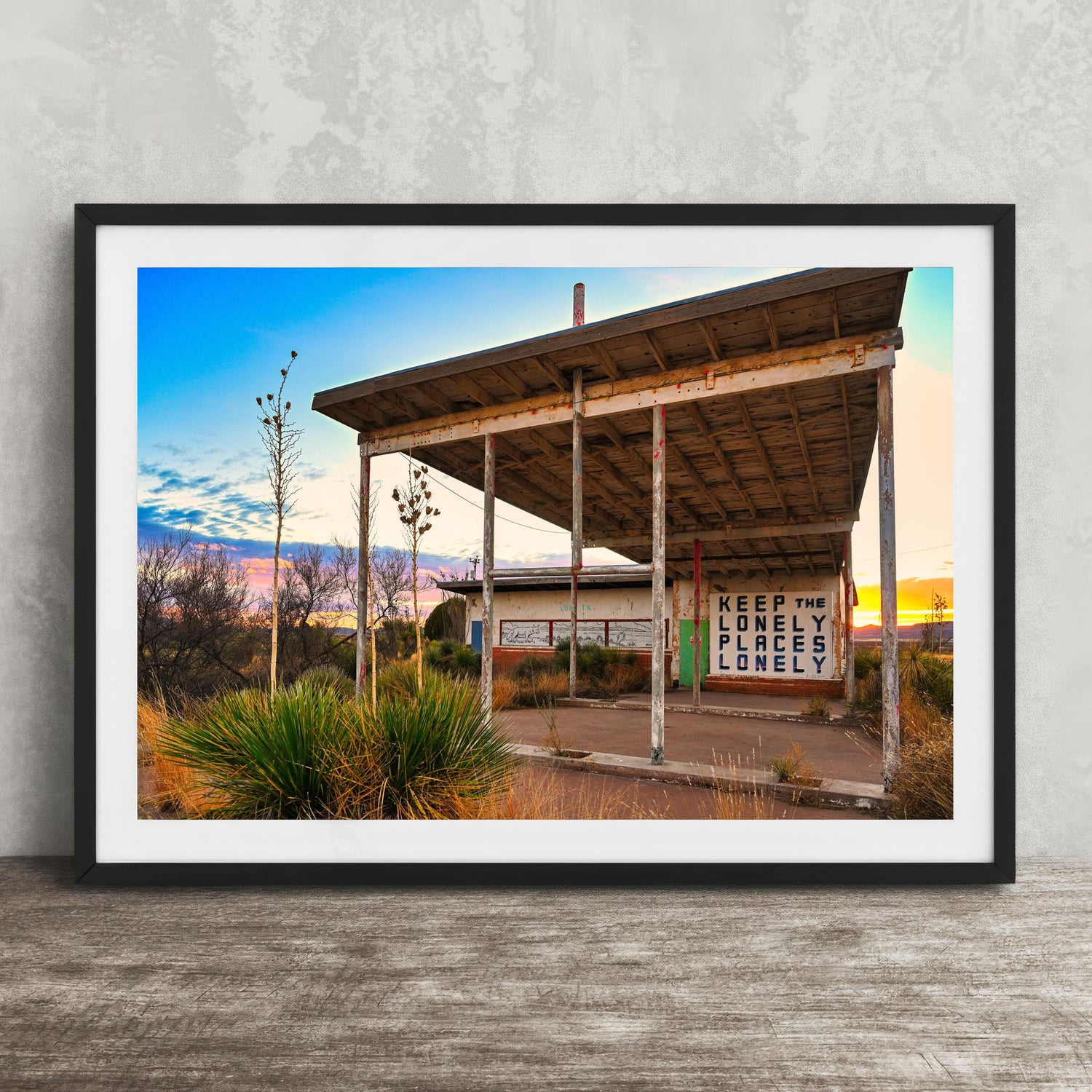 A framed photograph of a dilapidated wooden structure with a sign that reads "Keep the lonely lonely", set against a backdrop of a desert landscape with a colorful sunset sky.